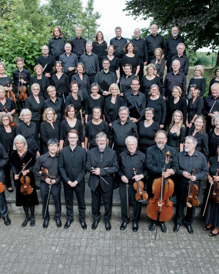 Ein großes Orchester steht draußen in formeller Kleidung, umgeben von grünen Bäumen. Sie halten verschiedene Musikinstrumente, darunter Geigen und Celli.
