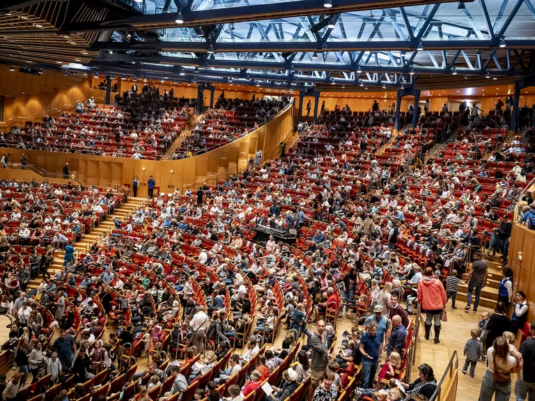 Kölner Philharmonie: A large, fully occupied auditorium featuring modern architectural elements. The audience is seated densely on red chairs.