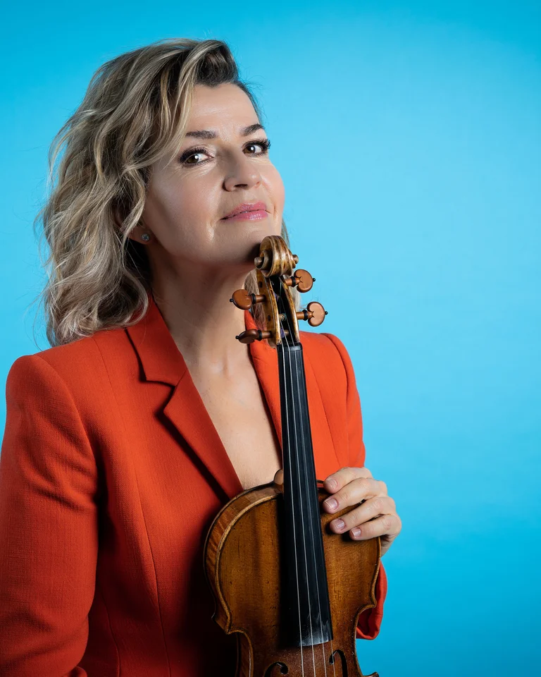 Woman in a red blazer holding a violin against a blue background. She appears confident and focused, with a slight smile.