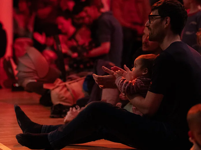 Menschen sitzen auf dem Boden eines dunklen Raumes, um eine Performance zu genießen. Der Raum ist in rotes Licht getaucht.