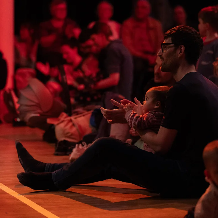 Menschen sitzen auf dem Boden eines dunklen Raumes, um eine Performance zu genießen. Der Raum ist in rotes Licht getaucht.