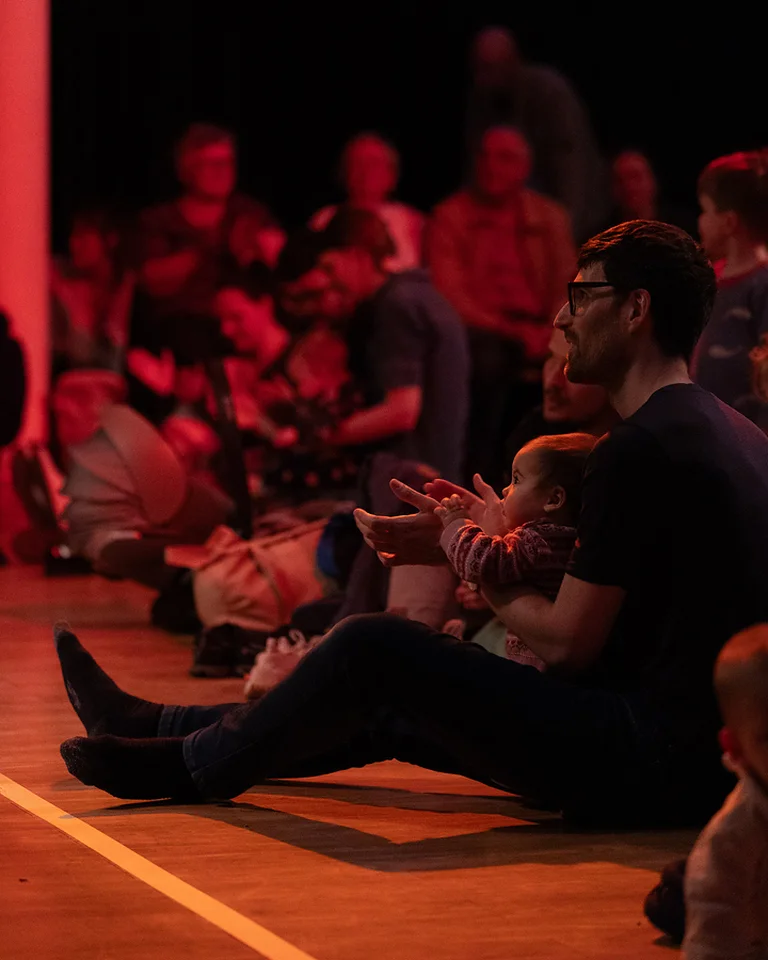 People seated on the floor in a dimly lit room, enjoying a performance. The room is illuminated by red light.