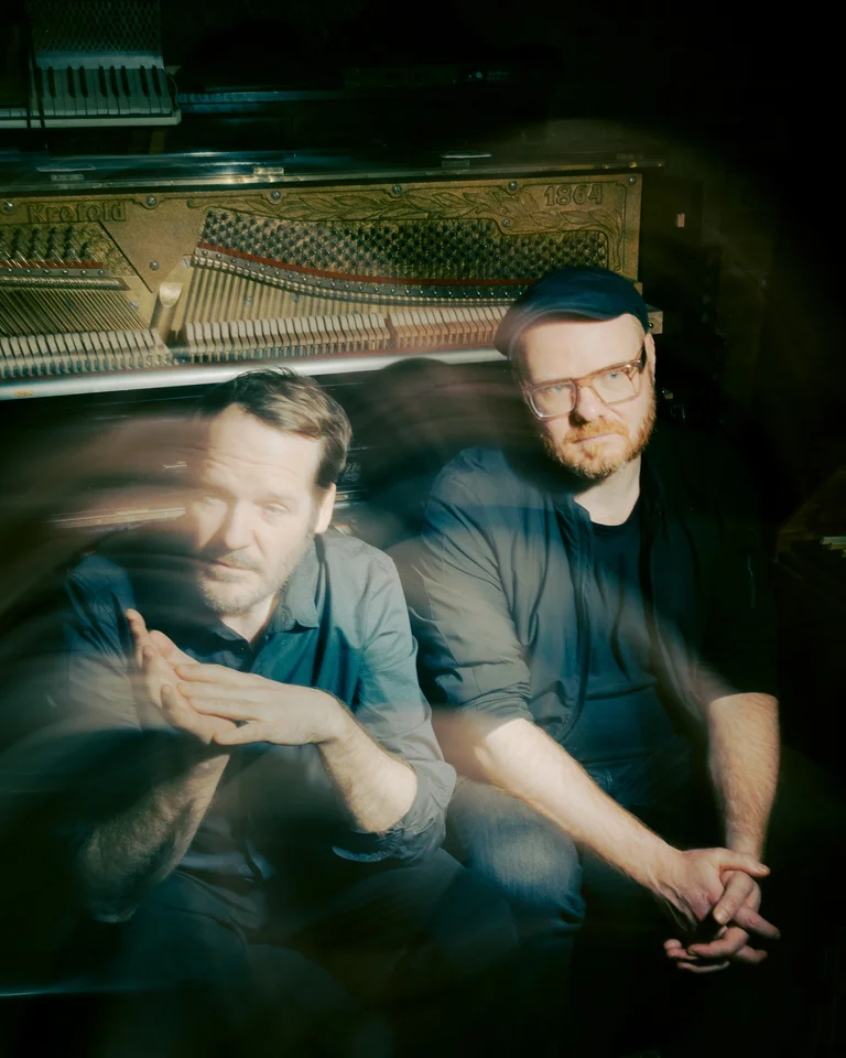 Zwei Männer sitzen vor einem verschwommenen Hintergrund mit einem Klavier. Atmosphärisches Bild mit einem geheimnisvollen Lichtspiel.