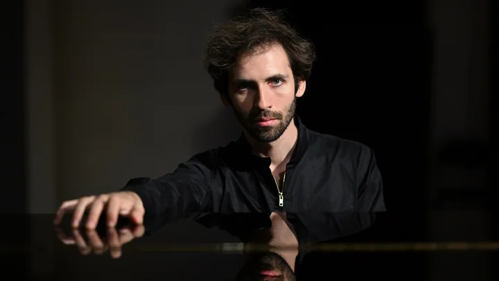 Ein Mann (Alexandre Kantorow) mit dunklem Haar und Bart sitzt an einem Klavier, wobei sein reflektiertes Bild auf der Oberfläche sichtbar ist.