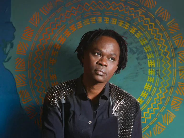 Eine Person in dunkler Kleidung steht vor einem farbenfrohen, kunstvollen Hintergrund mit spiralförmigen Mustern in Orange und Blau.