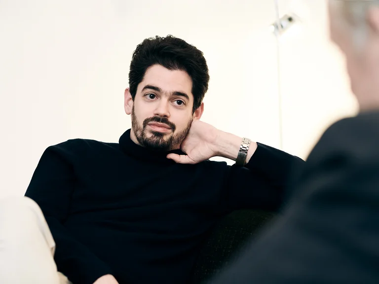 Ein Mann mit Bart in einem schwarzen Rollkragenpullover sitzt nachdenklich und entspannt, während er einen älteren Herrn ansieht.