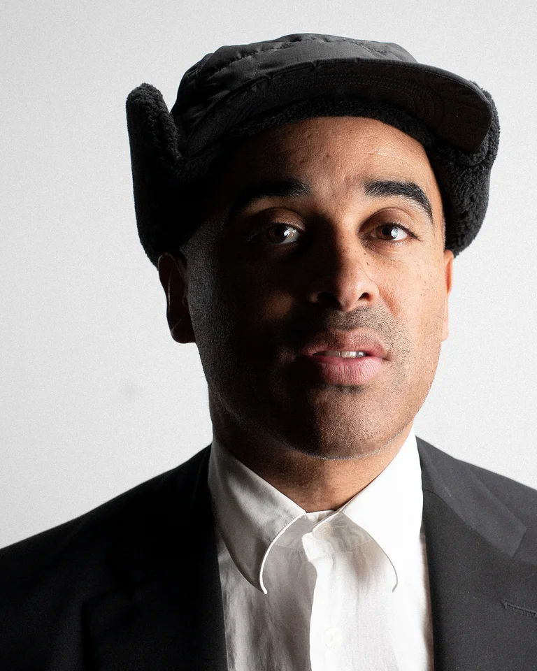 Patrice  wearing a black hat and suit against a light background, looking seriously at the camera.