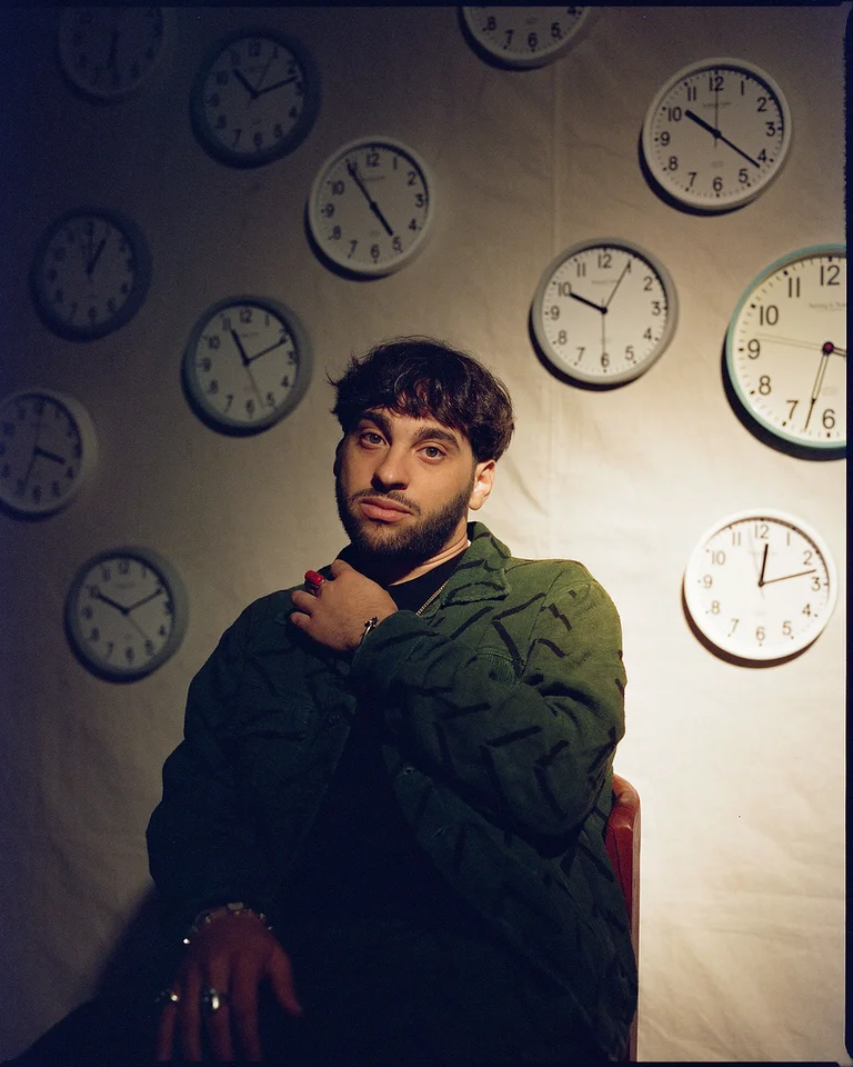 Ein Mann (Samora Pinderhughes) sitzt vor einer Wand mit vielen analogen Uhren. Er trägt einen dunkelgrünen Pullover und hat einen nachdenklichen Ausdruck.