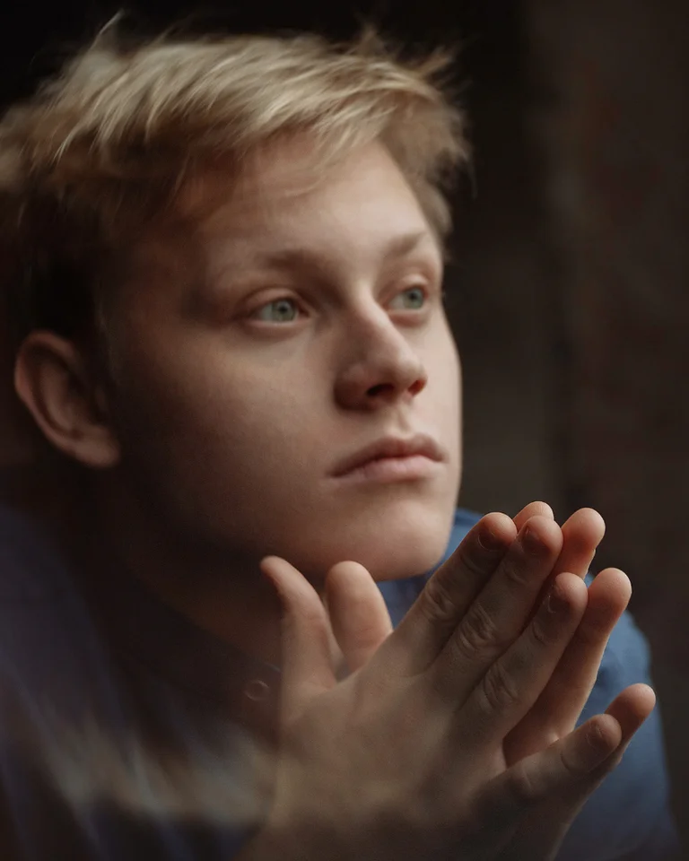 A young person looks thoughtfully out of a window. Their hands are clasped, expressing contemplation or anticipation.
