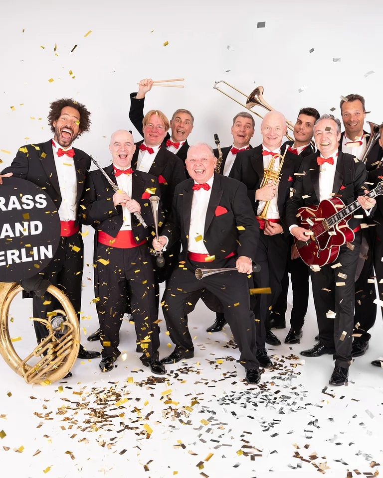 Eine fröhliche Blaskapelle in eleganten Anzügen, umgeben von Konfetti. Ein Musiker hält ein Schild mit "Brass Band Berlin".