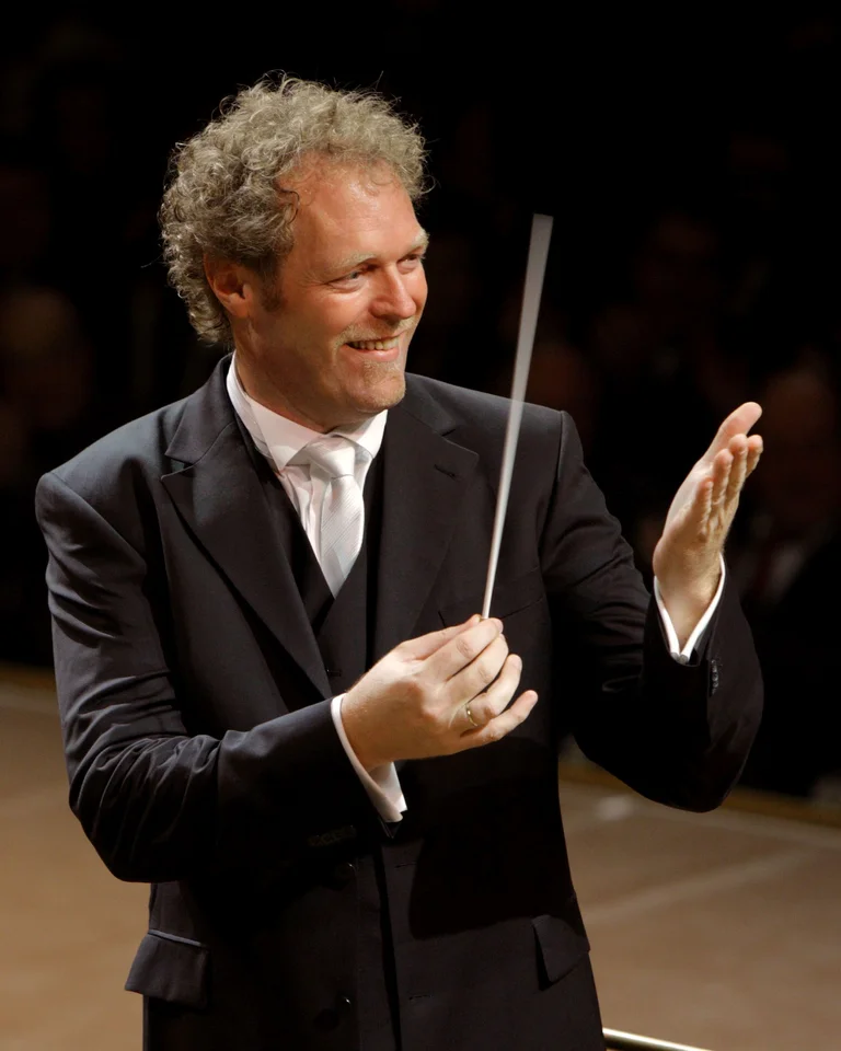 Ein Dirigent in einem schwarzen Anzug dirigiert ein Orchester. Er hält den Taktstock und spielt in einem Konzertsaal.