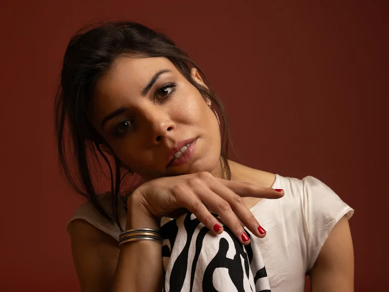 Eine Frau mit dunklem Haar, die ihren Kopf leicht geneigt hat. Sie trägt ein weißes Shirt und hat einen nachdenklichen Blick.