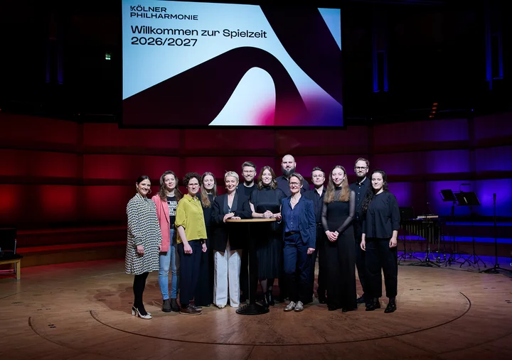 A group of people stands on a stage. In the background, a screen displays "Welcome to the 2026/2027 Season."