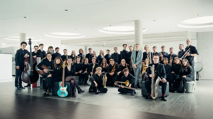 The members of the Bundesjazzorchester, dressed in black and holding various instruments, pose in a modern, light-filled room.