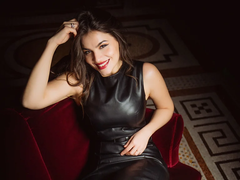 Eine Frau (Francesca Pia Vitale) in einem eleganten schwarzen Kleid sitzt lächelnd auf einem roten Sofa, umgeben von kunstvollem Fliesenboden.