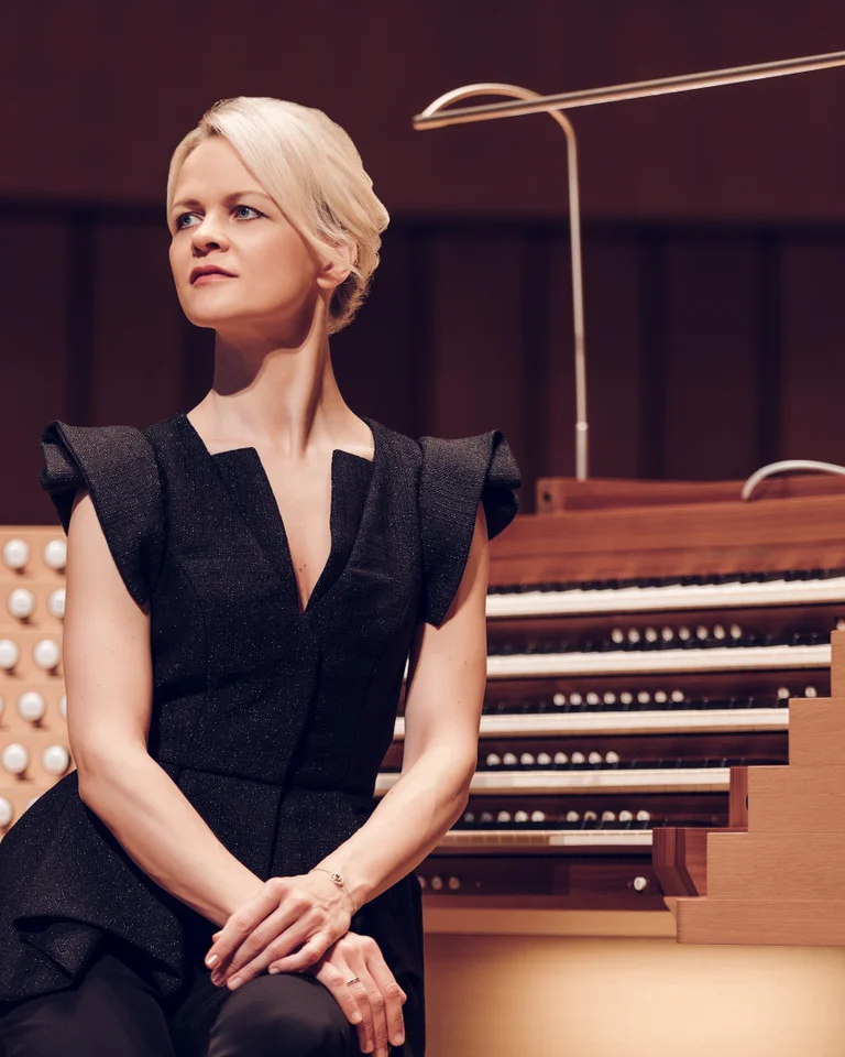 A woman in elegant attire stands beside a large organ in a concert hall. She gazes thoughtfully to the side.