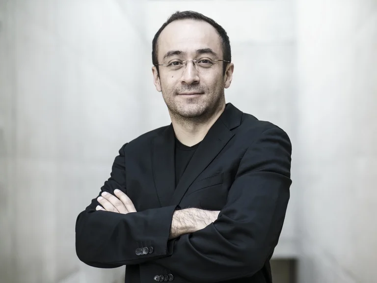 Mann mit verschränkten Armen im schwarzen Blazer (Riccardo Minasi), lächelnd; steht in einem hellen, eleganten Raum. Neutrales, selbstbewusstes Auftreten.