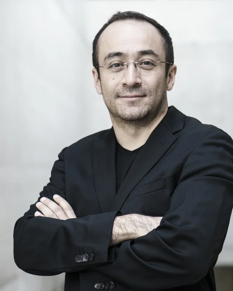 Mann mit verschränkten Armen im schwarzen Blazer (Riccardo Minasi), lächelnd; steht in einem hellen, eleganten Raum. Neutrales, selbstbewusstes Auftreten.