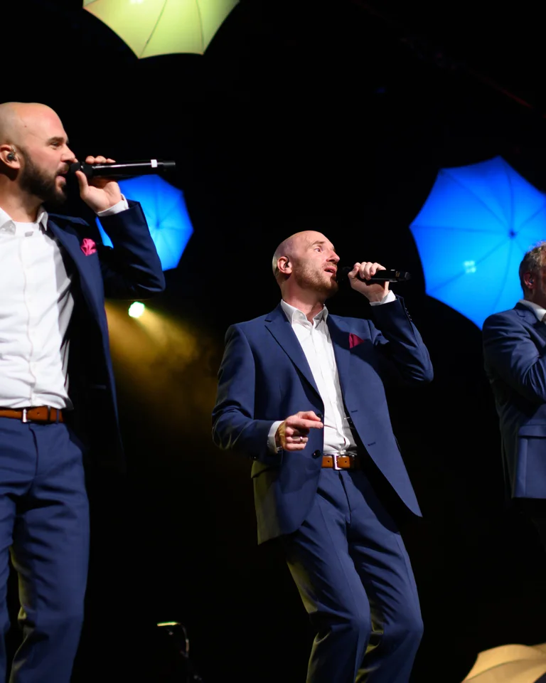 Fünf Männer in blauen Anzügen singen auf einer Bühne. Im Hintergrund leuchten bunte Bühnenscheinwerfer auf.