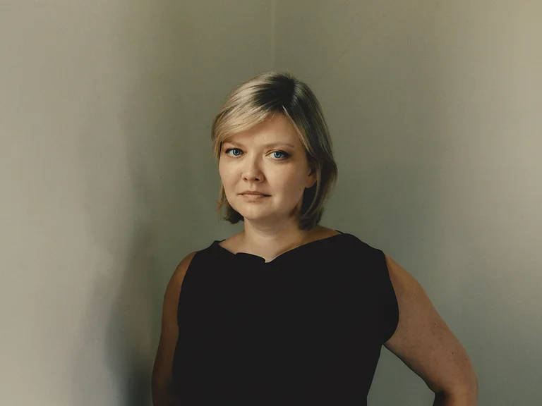 Eine Frau (Alina Ibragimova) mit kurzen blonden Haaren steht in einem dunklen Kleid vor einem neutralen Hintergrund.