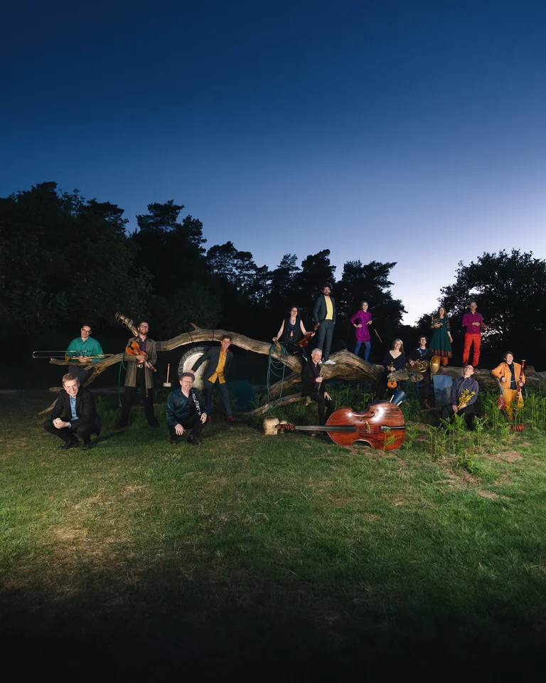 Eine Gruppe von Menschen (Ensemble Musikfabrik) posiert im Freien bei Dämmerung. Sie stehen und sitzen auf einem Baumstamm. Ein Kontrabass liegt im Gras.
