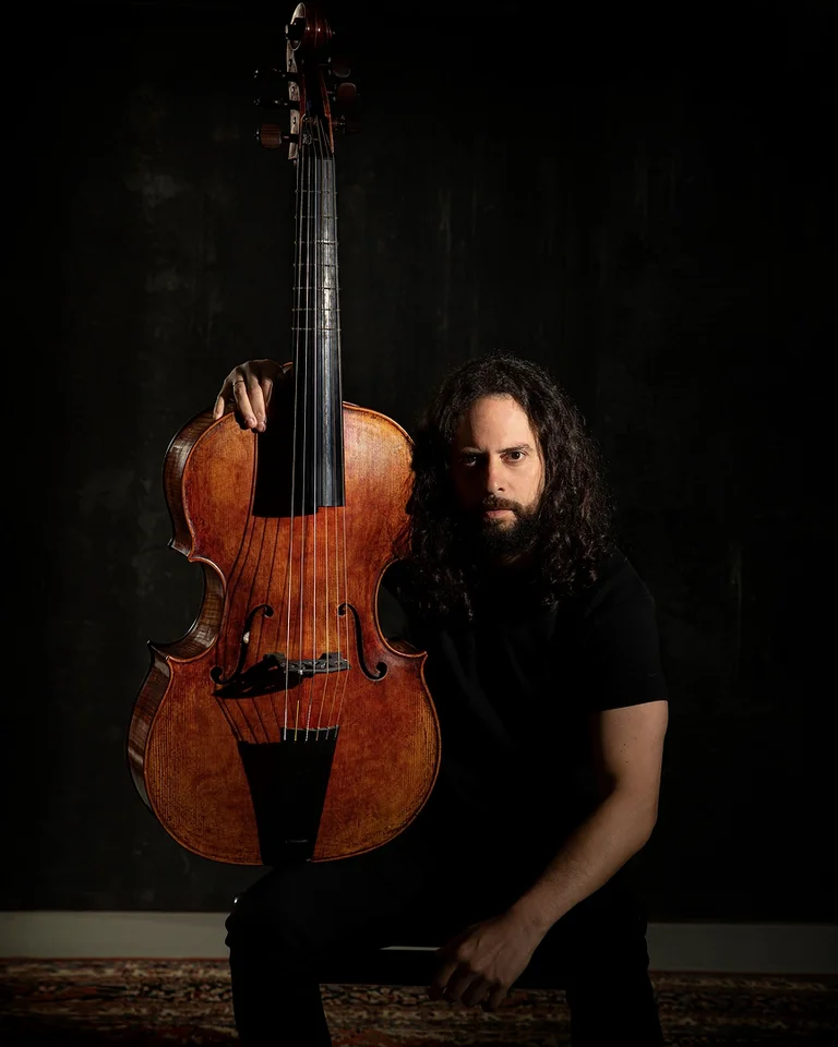 Ein Mann mit langen Haaren kniet neben einem großen, klassischen Kontrabass in einem dunklen Raum.
