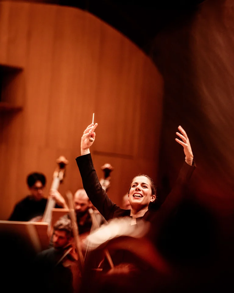 Dirigentin mit erhobenen Händen leitet ein Orchester. Der Konzertsaal ist warm beleuchtet, Musiker spielen verschiedene Streichinstrumente.