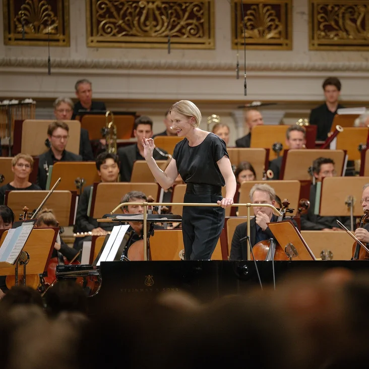 Eine Dirigentin leitet das Orchester (Joana Mallwitz mit Konzerthausorchester Berlin) in einem Konzertsaal. Musiker mit verschiedenen Instrumenten sind im Hintergrund zu sehen.