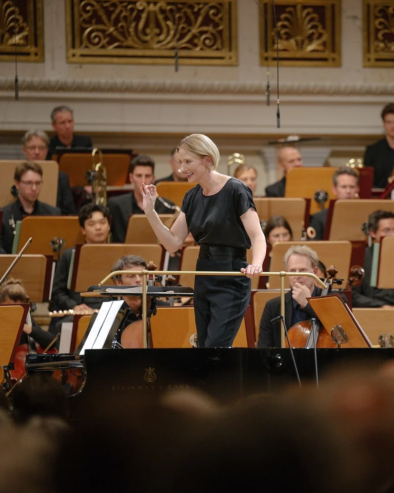 Eine Dirigentin leitet das Orchester (Joana Mallwitz mit Konzerthausorchester Berlin) in einem Konzertsaal. Musiker mit verschiedenen Instrumenten sind im Hintergrund zu sehen.