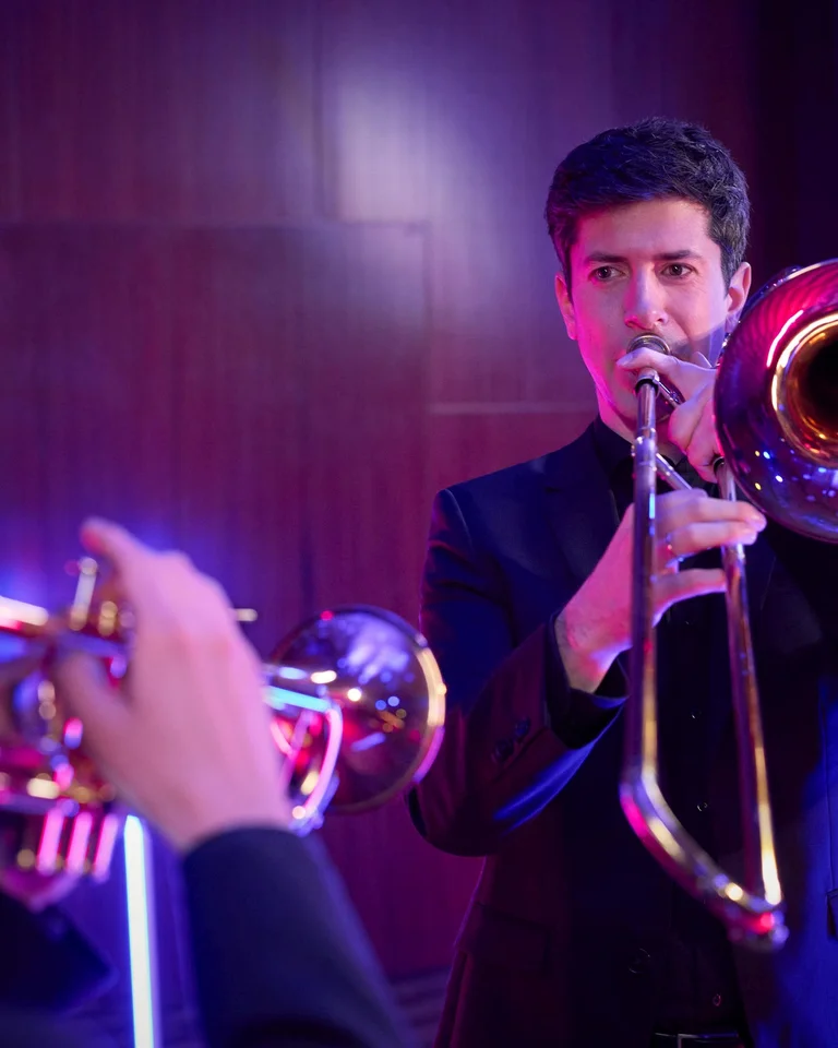 Ein Musiker spielt eine Posaune in einem modernen, beleuchteten Raum. Im Vordergrund ist ein Trompeter unscharf zu sehen.