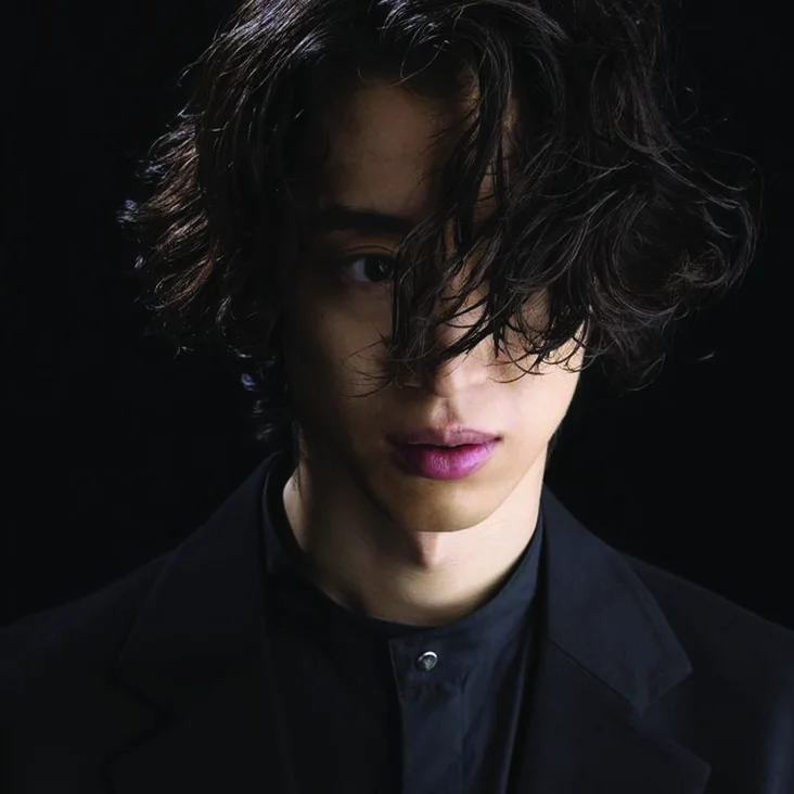 Ein Mensch mit welligem dunklem Haar trägt einen schwarzen Anzug und Hemd. Der Hintergrund ist schwarz, und das Licht ist subtil.