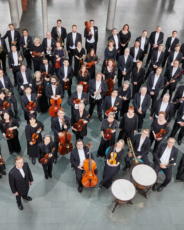 Ein großes Orchester in formeller Kleidung steht zusammen. Instrumente wie Streicher und Blasinstrumente sind sichtbar. Elegante Atmosphäre.