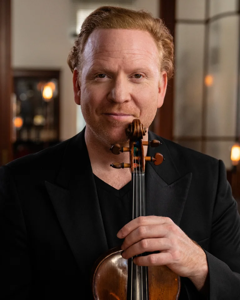 A man with curly hair holding a violin in a softly lit room with large windows and warm ambiance.