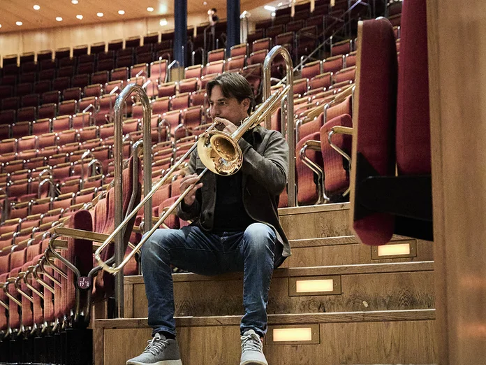 Person sitzt im leeren Konzertsaal auf Stufen und spielt Posaune, umgeben von leeren, gepolsterten Sitzreihen in der Kölner Philharmonie.
