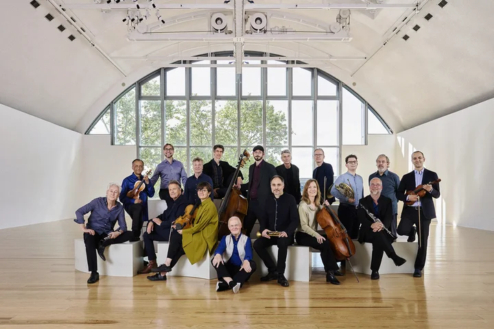 The members of Ensemble Modern pose standing and sitting in an elegant room with a large window and vaulted ceiling. Some of them are holding their instruments.
