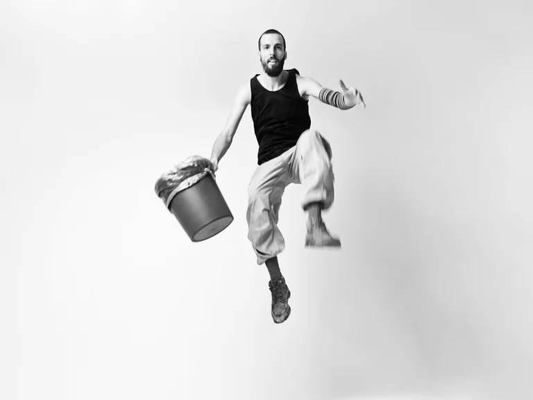 A man (Dominik Schad) in a sleeveless black shirt jumps into the air holding a bucket. Minimalist background.