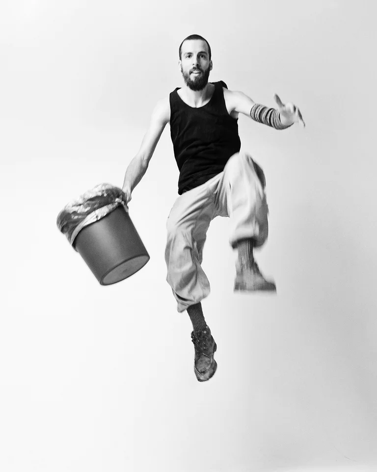 Ein Mann (Dominik Schad) in einem ärmellosen schwarzen Hemd springt in die Luft und hält einen Eimer. Minimalistischer Hintergrund.