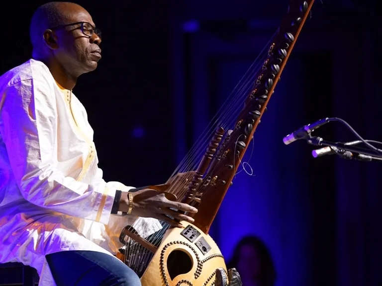 Ein Musiker (Sissoko Ballaké) spielt Kora, ein traditionelles westafrikanisches Saiteninstrument, auf einer Bühne in blauer Beleuchtung.