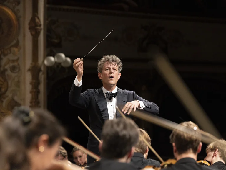 Der Dirigent Philipp von Steinaecker in eleganter Abendgarderobe leitet das Mahler Academy Orchestra während eines Konzertes, mit erhobenem Taktstock.
