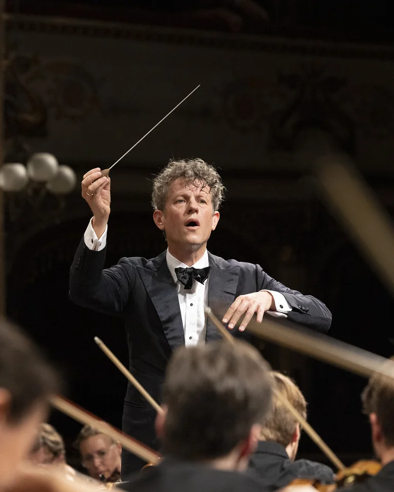 Der Dirigent Philipp von Steinaecker in eleganter Abendgarderobe leitet das Mahler Academy Orchestra während eines Konzertes, mit erhobenem Taktstock.