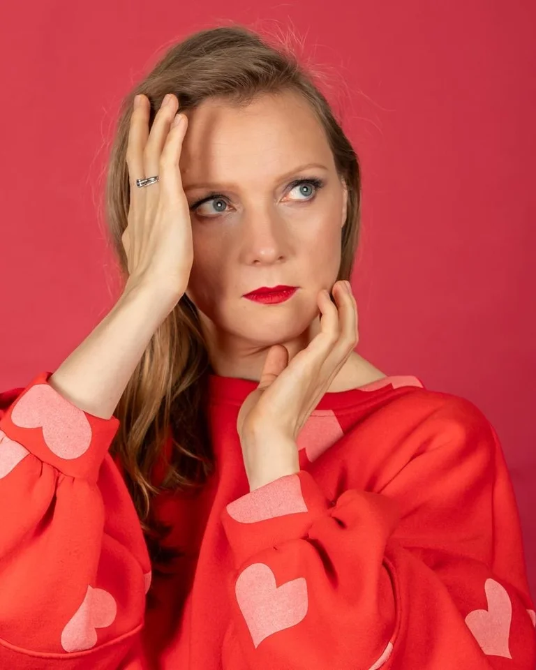 Frau (Olga Pashchenko) in rotem Pullover mit Herzmotiven posiert vor rotem Hintergrund, hält eine Hand an ihr Gesicht.