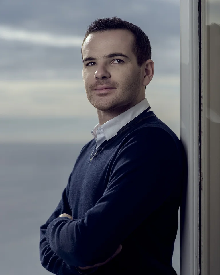 Ein Mann (Lionel Bringuier) steht vor einem Fenster mit Blick aufs Meer. Er trägt einen Pullover und blickt nachdenklich zur Seite.