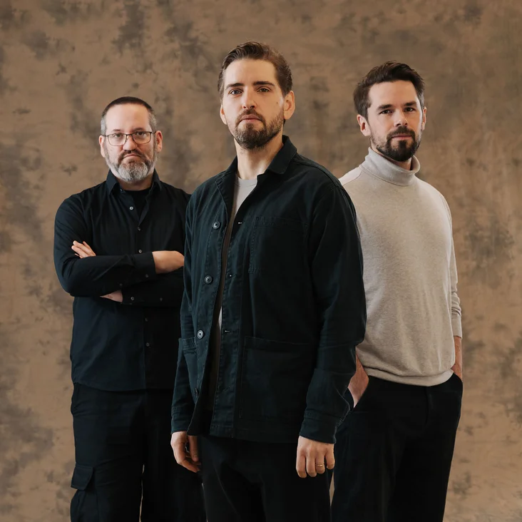 Drei Männer (Pablo Held Trio) stehen vor einem neutralen Hintergrund. Sie tragen lässige Kleidung und blicken ernst in die Kamera.