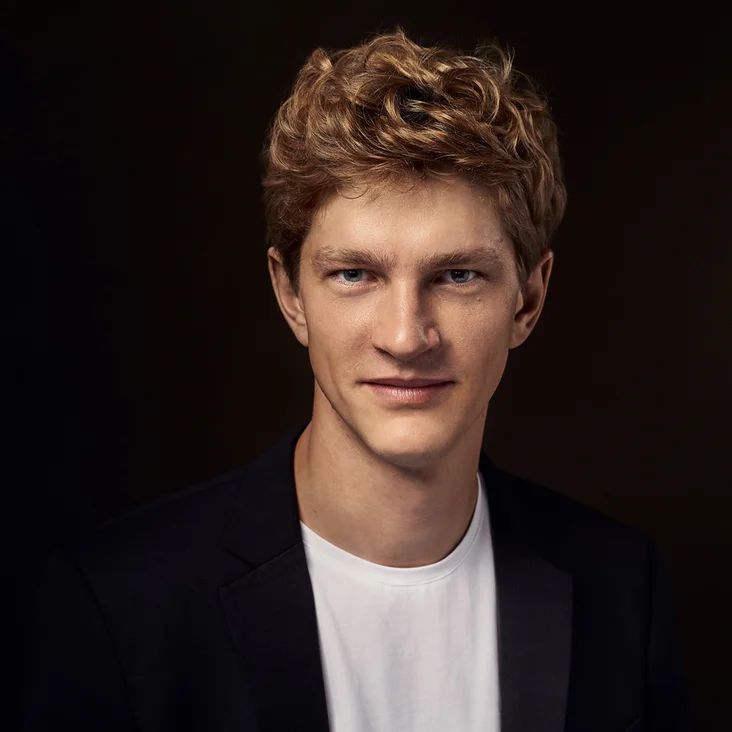 Porträt eines jungen Mannes mit lockigem Haar (Jan Lisiecki). Er trägt ein weißes T-Shirt und einen dunklen Blazer vor dunklem Hintergrund.