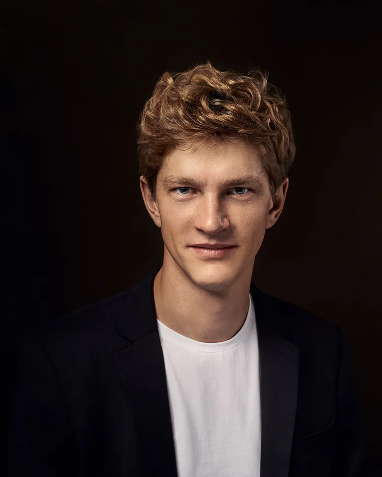 Porträt eines jungen Mannes mit lockigem Haar (Jan Lisiecki). Er trägt ein weißes T-Shirt und einen dunklen Blazer vor dunklem Hintergrund.