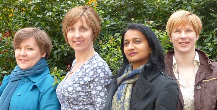 The four members of Synergy Vocals stand side by side and look into the camera. They are dressed in casual clothes and photographed outdoors.
