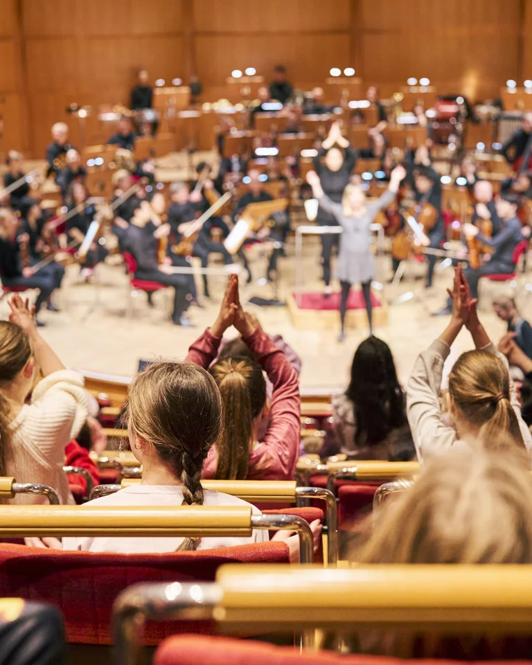 Ein Orchester spielt auf der Bühne vor einem Publikum. Kinder im Vordergrund klatschen begeistert. Der Konzertsaal ist modern gestaltet.