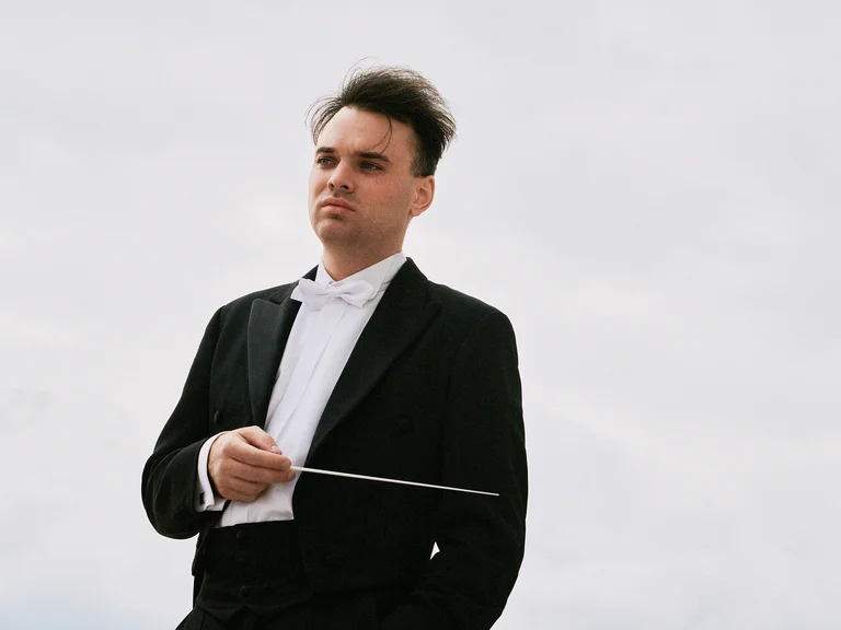 Ein Dirigent (Martin Rajna) in schwarzem Anzug und weißem Hemd hält einen Taktstock, vor einem bewölkten Himmel stehend.