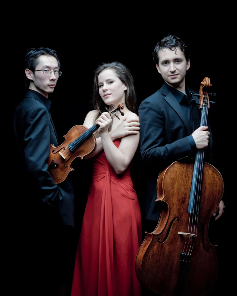 Drei Musiker posieren vor schwarzem Hintergrund. Ein Mann mit Violine, eine Frau in rotem Kleid und ein Mann mit Cello.