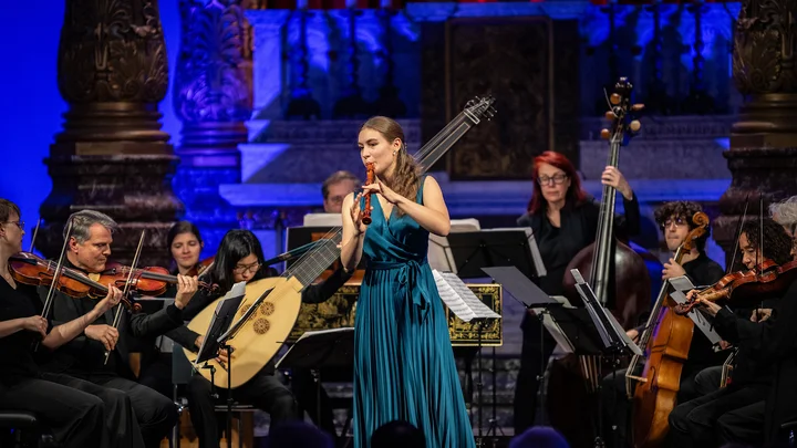 Eine Musikerin in einem blauen Kleid spielt eine Flöte vor einem Orchester mit Violen, Celli und einer Laute (Lucie Horsch mit dem Orchestra of the Eighteenth Century).